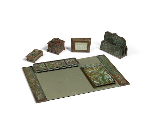 Tiffany Studios (1899-1930) Seven Piece Pine Needle Desk Setcirca 1910patinated bronze, Favrile glass, comprising calendar frame, letter holder, pair of blotter ends and pad, inkwell, stamp holder, pen tray and notepad holder, each element stamped 'TIFFANY STUDIOS NEW YORK' with various numbers, inkwell apparently unnumberedheight of letter holder 5 1/4in (13.3cm); width 6 1/4in (15.8cm); depth 2 3/4in (6.9cm)